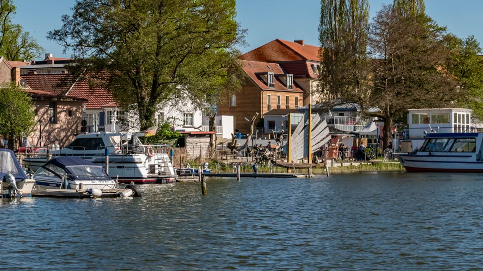 Riverside marina with several small boats and a larger cabin cruiser moored along wooden docks on calm water. Behind the docks are leafy trees and a row of low buildings with red-tiled roofs and light-colored facades.