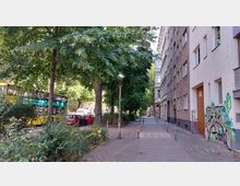 Straßenszene in einer Stadt mit einem schmalen Gehweg, der von hohen Wohngebäuden und Bäumen gesäumt ist. Links im Bild stehen geparkte Autos und ein gelber Doppeldeckerbus hinter einer kleinen Grünanlage, während die Gebäude rechts Fassaden mit Graffiti zeigen.