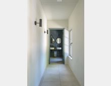 Schmaler, moderner Flur mit hellen Wänden und Bodenfliesen, der zu einem Badezimmer mit zwei Rundspiegeln, Waschbecken und dunklen Wandfliesen führt. Rechts hängt ein Fenster mit weißem Rahmen, und links sind schwarze Wandlampen angebracht.