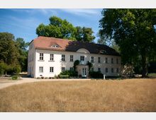 Zweistöckiges, historisch wirkendes Gebäude mit hellem Putz und einem roten Satteldach, umgeben von Bäumen und einer trockenen Wiese im Vordergrund. Der Eingang ist von Rankpflanzen umgeben und führt zu einem kleinen Balkon über der Tür.