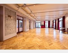 Ein geräumiger Veranstaltungsraum mit hellem Fischgrätenparkett, freiliegenden Ziegelwänden und großen Glas-Doppeltüren. Der Saal ist mit roten Vorhängen an den Fenstern, sichtbaren Deckenrohren und moderner Beleuchtung ausgestattet.