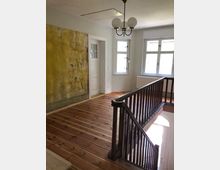 Ein helles Treppenhaus mit Holzboden und einem dunklen Holzgeländer. An der linken Wand befindet sich eine gelbe, leicht abblätternde Fläche, daneben eine weiße Tür mit Glaseinsatz; rechts sind drei Fenster, die Tageslicht einlassen.