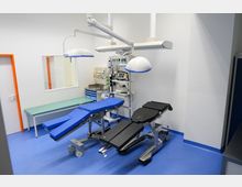 Ein steriler Operationsraum mit blauen Bodenfliesen, zwei verstellbaren OP-Tischen (einer mit blauem, der andere mit schwarzem Polster) und zwei Deckenleuchten. Im Hintergrund befinden sich medizinische Geräte, ein grünes Untersuchungslager und ein Fenster in die angrenzende Wand.