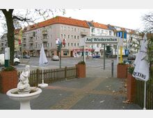 Straßenszene mit einem kleinen Platz im Vordergrund, auf dem eine weiße Statue auf einem Brunnen steht, umgeben von niedrigen Zäunen und Backsteinkübeln mit Pflanzen. Im Hintergrund ist eine Kreuzung mit Wohn- und Geschäftshäusern sowie einem Schild mit der Aufschrift „Auf Wiedersehen“ sichtbar.