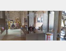 Offener Wohnbereich im Industriestil mit Sichtziegelwänden, einem modernen Kamin mit Holzlager, einer Kücheninsel aus Metall und modernen Designdetails. Im Hintergrund Glaselemente als Raumteiler, Kunstwerke an den Wänden und eine offene Metalltreppe.