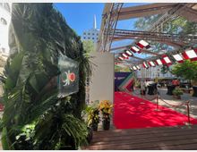 Ein Außenbereich mit einem roten Teppich, umgeben von Absperrungen und dekoriert mit Pflanzen. Eine überdachte Konstruktion aus Metall und Glas mit Beleuchtung überspannt den Bereich, im Hintergrund ist ein modernes Gebäude mit Spitze sichtbar.