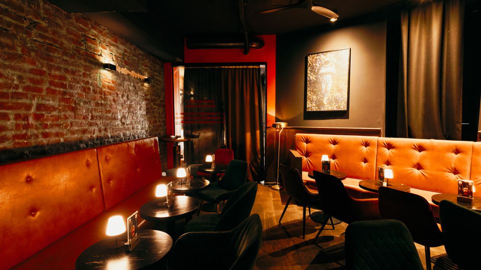 Innenraum einer gedimmten Bar-Lounge mit freiliegender Backsteinwand, gepolsterten orangefarbenen Sitzbänken und kleinen runden Tischen mit Tischlampen. Dunkle Decke mit Lüftungsrohren und Ventilator, schwarze Vorhänge sowie ein gerahmtes Bild an der Wand.