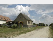 Ein schlichtes Einfamilienhaus mit Satteldach, verputzter Fassade und kleinen Fenstern, umgeben von einem Zaun in einer ländlichen Umgebung. Rechts davon verläuft ein unbefestigter Feldweg, im Hintergrund sind Bäume und eine offene Landschaft zu sehen.