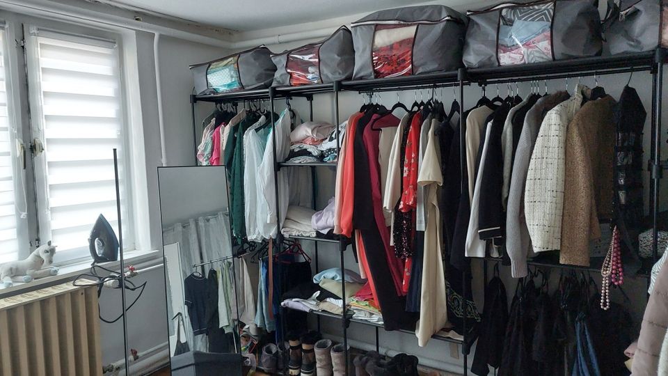 Kleiner Ankleide- bzw. Abstellraum mit einem offenen Kleiderregal aus Metall, an dem viele Jacken und Oberteile hängen, sowie Fächern mit gefalteter Kleidung und Stofftaschen oben. Links befindet sich ein Fenster mit Lamellenrollo, darunter ein Heizkörper; daneben steht ein Spiegel und ein Bügeleisen.