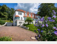 Weißes, zweigeschossiges Gebäude mit roten Ziegeldächern und Bogenfenstern, umgeben von einem Garten mit blühenden lila Blumen im Vordergrund. Ein gepflasterter Hof führt zum Eingangsbereich, der von einer kleinen Treppe flankiert wird.