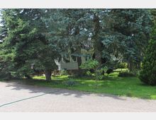 Ein Wohnhaus aus Klinkersteinen ist hinter großen Nadelbäumen erkennbar, die einen grünen, gepflegten Garten mit vereinzelten Sträuchern und Pflanzen umgeben. Im Vordergrund befindet sich ein gepflasterter Weg mit einem Wasserschlauch darauf.