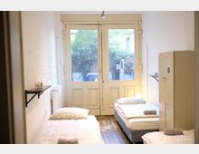 Ein einfacher Schlafraum mit drei Einzelbetten, weißer Bettwäsche und kleinen Handtüchern darauf. Im Hintergrund befinden sich große Doppeltüren mit Glas, die den Blick auf einen begrünten Innenhof freigeben; an den Wänden sind ein Regalbrett sowie ein Spind angebracht.