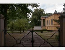 Gelber historischer Gebäudeflügel mit Säulenvorbau, umgeben von Bäumen und einer gepflegten Gartenanlage, gesehen durch ein schmiedeeisernes Tor.