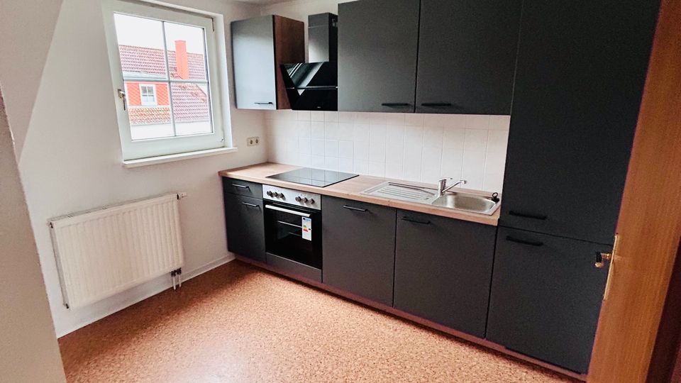 Helle, leere Küche mit schräger Decke, großem Fenster und Heizkörper unter dem Fenster. Entlang einer Wand verläuft eine moderne, dunkelgraue Einbauküche mit Herd, Dunstabzug, Spüle und weißem Fliesenspiegel.