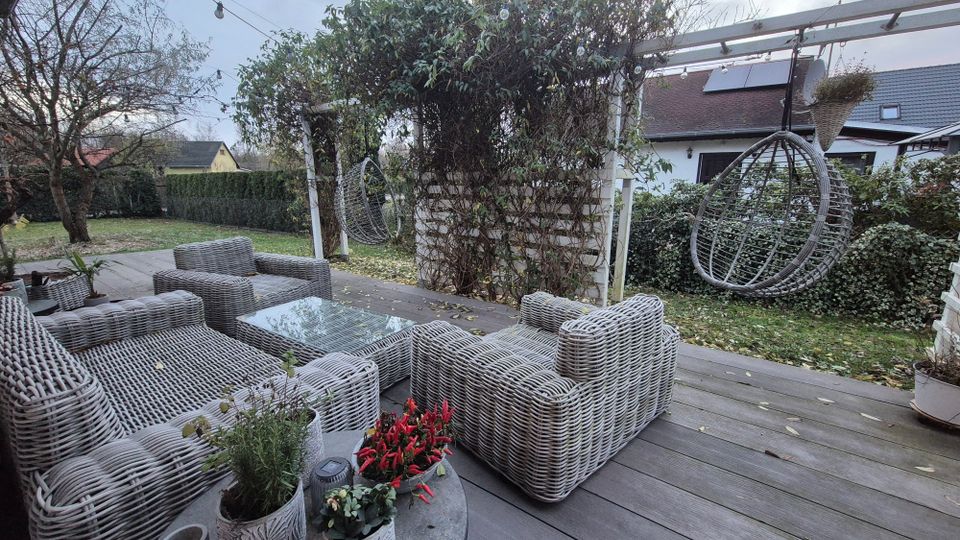 Terrasse mit grauen Rattanmöbeln, darunter Sessel und ein Tisch mit Glasplatte. Im Hintergrund befindet sich eine Pergola mit Kletterpflanzen, daneben ein hängender Rattansessel. Der Garten ist mit Rasen, Hecken und Bäumen gestaltet, während im Hintergrund Häuser und ein bewölkter Himmel zu sehen sind.