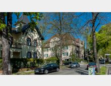 Straßenszene in einem Wohngebiet mit zwei größeren Häusern im traditionellen Stil, verputzten Fassaden und Satteldächern. Umgeben von Bäumen mit frischem Laub und parkenden Autos entlang der Straße.