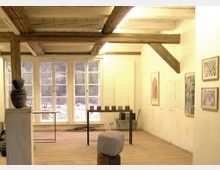 Ein heller Raum mit Holzbalkendecke und Holzboden, großen Sprossenfenstern, die eine winterliche Landschaft zeigen. Im Vordergrund stehen Skulpturen und ein Tisch mit kleinen Objekten, an den Wänden hängen Bilderrahmen.