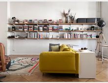 Modern living room with a mustard-yellow sofa facing a long wall of floating wooden shelves filled with books, records, speakers, and decorative vases on a white wall. A patterned area rug and a white side table sit on tan tile flooring, with a large black tripod floor lamp in the corner.