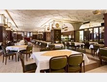 Ein geräumiges Restaurant mit klassischer Inneneinrichtung. Es verfügt über viele gedeckte Tische mit weißen Tischdecken, dunklen Holzstühlen und eine verzierte Decke mit geometrischem Muster.