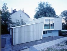Modernes Gebäude mit wellenförmiger Metallfassade und großen Glasfenstern, umgeben von Kiesflächen und Gehwegen. Im Hintergrund ist ein älteres Wohnhaus und dichter Baumbestand zu sehen.