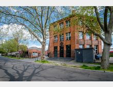 Backstein-Gebäude mit großen Fenstern und einem Treppenaufgang an der Vorderseite, umgeben von Bäumen mit frischem Laub. Der Standort befindet sich an einer ruhigen Straße mit Gehwegen und vereinzelten weiteren Backsteinbauten in der Umgebung.