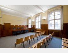 Ein heller, historischer Gerichtssaal mit hohen Decken, verzierten Stuckelementen und großen Fenstern, die Tageslicht einlassen. Der Raum ist mit hölzernen Wandpanelen, einer Richterbank mit Stühlen und Sitzreihen aus Holz für Zuschauer ausgestattet.