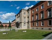 Historisches Gebäude mit rotbrauner und beiger Fassade, großen Fenstern und klassischen Architekturelementen, umgeben von einem gepflegten Rasen mit Steinbänken; im Hintergrund blauer Himmel mit weißen Wolken.