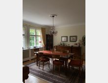 Esszimmer mit Holztisch und sechs gepolsterten Stühlen auf einem gemusterten Teppich. Im Hintergrund sind eine Kommode mit Getränken und Gläsern, ein Buffetschrank, zwei gerahmte Bilder an der Wand, ein Kronleuchter an der Decke sowie ein Fenster mit Vorhängen zu sehen.