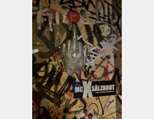 Nahaufnahme einer mit Graffiti, Kritzeleien und Aufklebern bedeckten Wand, in deren Mitte eine silberne Hand-Skulptur montiert ist. Unten ist ein rechteckiger Sticker mit der Aufschrift „MC SÄLZBROT“ zu sehen.