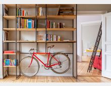 Heller Wohnraum mit großem, offenen Bücherregal aus Holz und schwarzem Metall, gefüllt mit Büchern und einigen Gegenständen; darunter steht ein rotes Fahrrad vor der Regalwand. Rechts führt eine offene Tür in ein Schlafzimmer mit Metallbett, daneben lehnt eine schwarze Leiter am Durchgang.