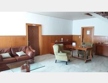 Ein Loungebereich mit Holzvertäfelung an den Wänden, zwei Holztüren und einem Boden aus polierten Fliesen. Im Vordergrund steht ein Glastisch mit einer Ledercouch und grünen Sesseln, daneben ein Schreibtisch mit Dekorationen.