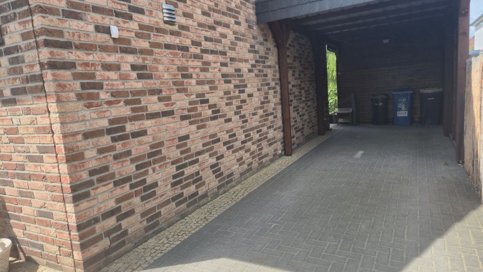 Überdachte Einfahrt neben einem Backsteinhaus mit gepflastertem Boden, Holzbalken und einem flachen, schindelgedeckten Dach. Am Ende des Carports stehen mehrere Mülltonnen, rechts begrenzt von einem Holzzaun.