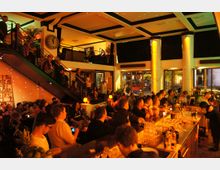 Belebte Bar mit moderner Einrichtung, großer Theke im Vordergrund, Sitz- und Stehbereich im Erdgeschoss sowie Galerieebene mit Treppe. Warmes Licht und dekorative Elemente schaffen eine gemütliche Atmosphäre.