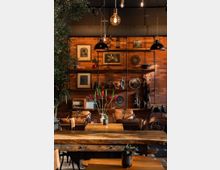 Ein Café mit rustikalem Design, geprägt von einer Backsteinwand mit dekorativen Regalen voller Retro-Gegenstände, Bildern und Pflanzen. Im Vordergrund stehen Holztische mit einfachen Dekoelementen, begleitet von braunen Ledersesseln.