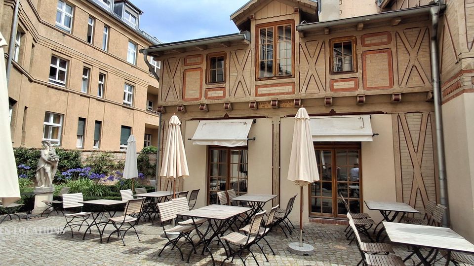 Innenhof mit Kopfsteinpflaster und einer Café-Terrasse mit Metalltischen, Stühlen und geschlossenen Sonnenschirmen. Im Hintergrund steht ein historisches Fachwerkgebäude mit großen Fenstern und Markisen, flankiert von weiteren Gebäudefassaden, Blumenbeeten und einer kleinen Statue.