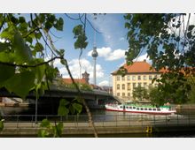 Blick auf die Spree in Berlin mit einem Ausflugsboot im Vordergrund, darüber eine Brücke. Im Hintergrund sind der Berliner Fernsehturm, ein historisches Gebäude mit rotem Dach und Teile des Nikolaiviertels zu sehen, eingerahmt von Laubzweigen.