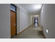 Langer, schlichter Flur mit hellen Wänden und glattem Bodenbelag, rechts an der Wand befinden sich schlichte Sitzbänke und Wandleuchten, links sind Holztüren mit Beschilderung. Am Ende des Flurs ist eine Glastür sichtbar.