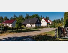 Eine ruhige Wohnsiedlung mit Einfamilienhäusern und gepflegten Vorgärten, umgeben von Bäumen. Die Straße ist unbefestigt, und einige Häuser haben Zäune aus Metall.