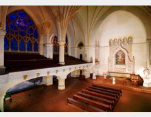 Innenraum einer historischen Kirche mit hohen gotischen Gewölbedecken, einer großen Buntglas-Rosette auf der linken Seite und einer Altarwand mit religiösem Gemälde und Statuen. Reihen von Holzbänken und eine große Empore prägen das Raumdesign.