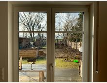 Blick durch eine Terrassentür auf einen Garten mit Rasen, Bäumen und Holzhochbeet im Vordergrund. Im Hintergrund stehen ein Schuppen und verschiedene Gartenutensilien, umgeben von einem Holzzaun.