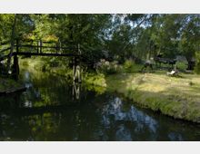 Holzbrücke über einen schmalen, von Bäumen umgebenen Kanal mit blühenden Pflanzen am Ufer und alter landwirtschaftlicher Ausrüstung im Hintergrund auf einer grasbewachsenen Fläche.