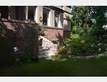 Backsteinhaus mit kleinem Garteneingang, dessen weiße Steintreppe zu einer Tür führt. Im Vordergrund ist ein gepflegter Garten mit Blumenbeeten, einem Rasen und dekorativen Pflanzgefäßen zu sehen.