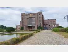 Backsteinbau mit symmetrischer Fassade und großen Fensterreihen, flankiert von gepflegten Büschen und einem Wasserspiegel im Vordergrund, mit einem gepflasterten Weg, der zum Eingang führt. Im Hintergrund befinden sich grüne Wiesen und Bäume unter einem teilweise bewölkten Himmel.