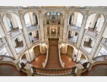 Innenansicht eines historischen Gebäudes mit einer großen, geschwungenen Haupttreppe aus Stein, umgeben von mehreren Etagen mit Arkadengängen und dekorativen Balustraden. Die Architektur ist geprägt von symmetrischen Bögen, Säulen und kunstvoll gestalteten Details.