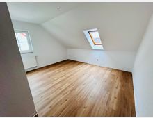 Leerer Dachgeschossraum mit schrägen weißen Wänden und hellem Holzfußboden. Ein normales Fenster mit Heizkörper darunter sowie ein Dachfenster sorgen für Tageslicht.