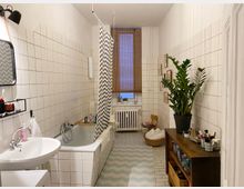 Schmales Badezimmer mit weißen Wandfliesen und einer Badewanne mit grauem Duschvorhang. Im Raum befinden sich ein Waschbecken mit Spiegel, ein Regal mit einer großen Zimmerpflanze, ein Heizkörper unter einem Fenster mit Holzrollo und weitere Dekorationselemente wie gerahmte Bilder und ein Teppich mit Zickzack-Muster.