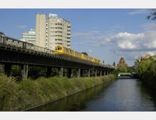 Gelbe U-Bahn auf einer erhöhten Bahntrasse über einem Kanal, flankiert von dichter Vegetation. Im Hintergrund sind moderne Wohnhäuser und eine Kirche mit rotem Backstein und Kuppeldach zu sehen, bei leicht bewölktem Himmel.