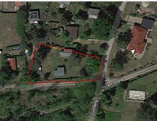 Satellitenansicht eines ländlichen Anwesens, das von einem roten Umriss markiert ist. Das Grundstück enthält eine graue Struktur und ist von Bäumen und angrenzenden Grundstücken umgeben. Zwei Straßen, "Zum Berg" und "Am Strang," grenzen an die Fläche.