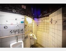 Eine öffentliche Herrentoilette mit gefliesten Wänden in Weiß, die mit Graffiti und Aufklebern bedeckt sind. Im Vordergrund sind zwei Urinale zu sehen, während grelle Beleuchtung und violette Akzente die düstere Atmosphäre betonen.