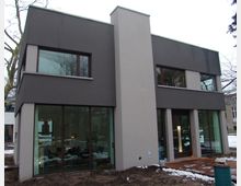 Zweistöckiges modernes Wohnhaus mit Flachdach und klaren Linien, gestaltet in hellen und dunkelgrauen Fassadenfarben. Die großen Glasfensterfronten bieten Einblicke in das Interieur; umliegendes Grundstück ist teilweise schneebedeckt.